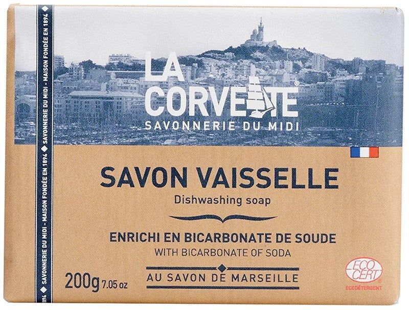 La Corvette Astianpesusaippua; La Corvette-astianpesusaippuapala, 200 g, esillä Marseillen kaupunkimaiseman edessä; pakkaus suorakaiteen muotoinen mattaruskeaa paperia sinisin tehostein; ranskan- ja englanninkielistä tekstiä lihavoidulla groteskikirjasimella, näkyvissä ECOCERT-logo; ei ihmisiä tai käsiä kuvassa; tuote keskitettynä suorassa kuvassa.