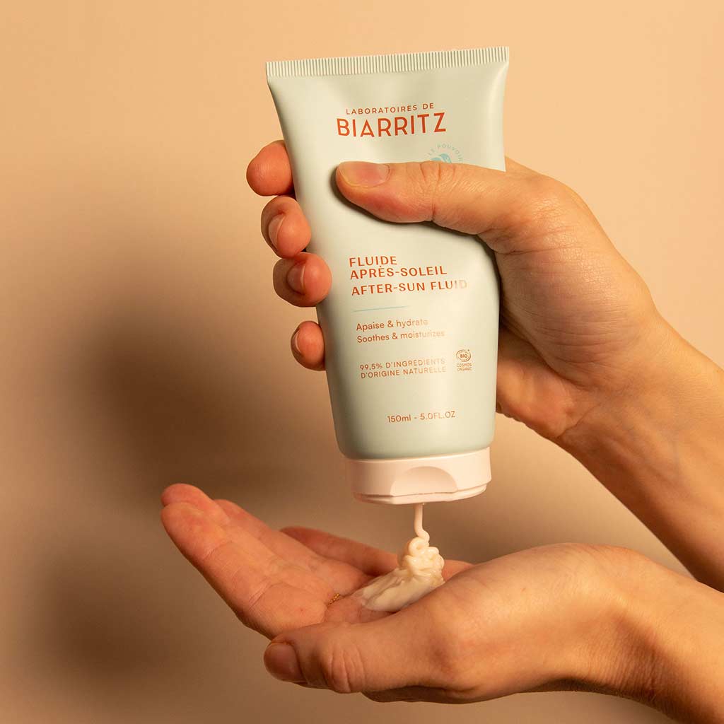 Laboratoires de Biarritz After-Sun Fluid: henkilö annostelee valkoista voidetta tuubista tekstillä "Laboratoires de Biarritz After-Sun Fluid" kämmenelleen beigeä taustaa vasten, korostaen rauhoittavaa aurinko-jälkihoitoa; valkoinen muovituubi on mattapintainen; etiketissä musta groteskiteksti; tuubi on suorakulmainen pyöristetyin reunoin ja läppäkorkilla; tausta on tasaisen beige ilman muita esineitä; lähikuva keskittyy käteen ja tuotteen annosteluhetkeen.