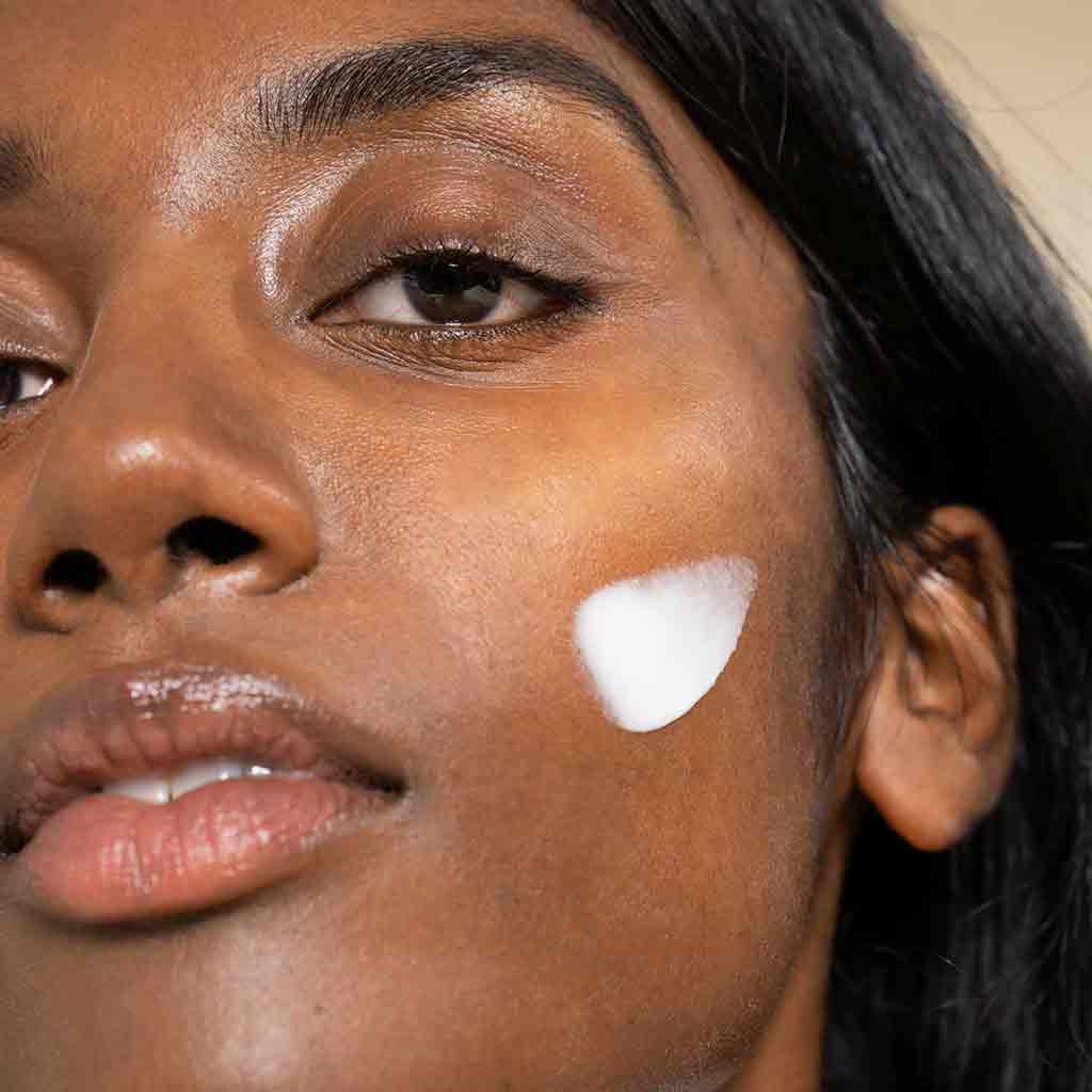 Laboratoires de Biarritz Anti Dark Spot Night Cream: lähikuva tummaihoisesta naisesta, jonka poskella on valkoista voidetta; katse on suunnattu kameraan neutraalilla ilmeellä, kuva korostaa ihonhoitoa; tuotepakkausta tai etikettiä ei näy; tausta on vaalea ja tasaisesti valaistu, kädet eivät ole näkyvissä.