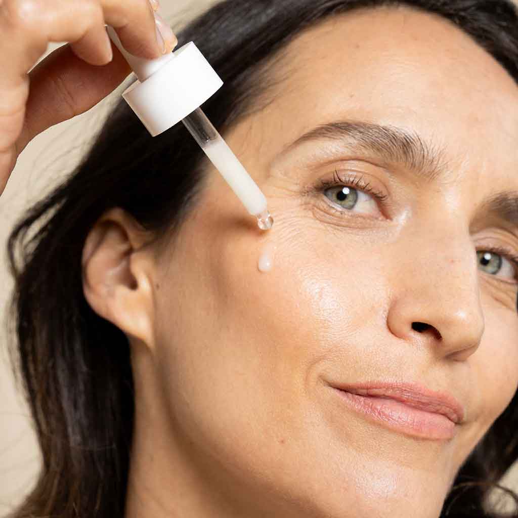 Laboratoires de Biarritz Anti Dark Spot Serum: nainen levittää kosteuttavaa kasvoseerumia poskelleen pipetin avulla, iho vaikuttaa sileältä ja hän katsoo kameraan pieni hymy huulillaan; pipetti on läpinäkyvä ja kuuluu pieneen, läpikuultavaan pulloon, jossa on kirkasta nestettä; pakkauksessa ei näy tekstiä; tausta on vaalea, kuvassa näkyy vain käsi ja kasvot.