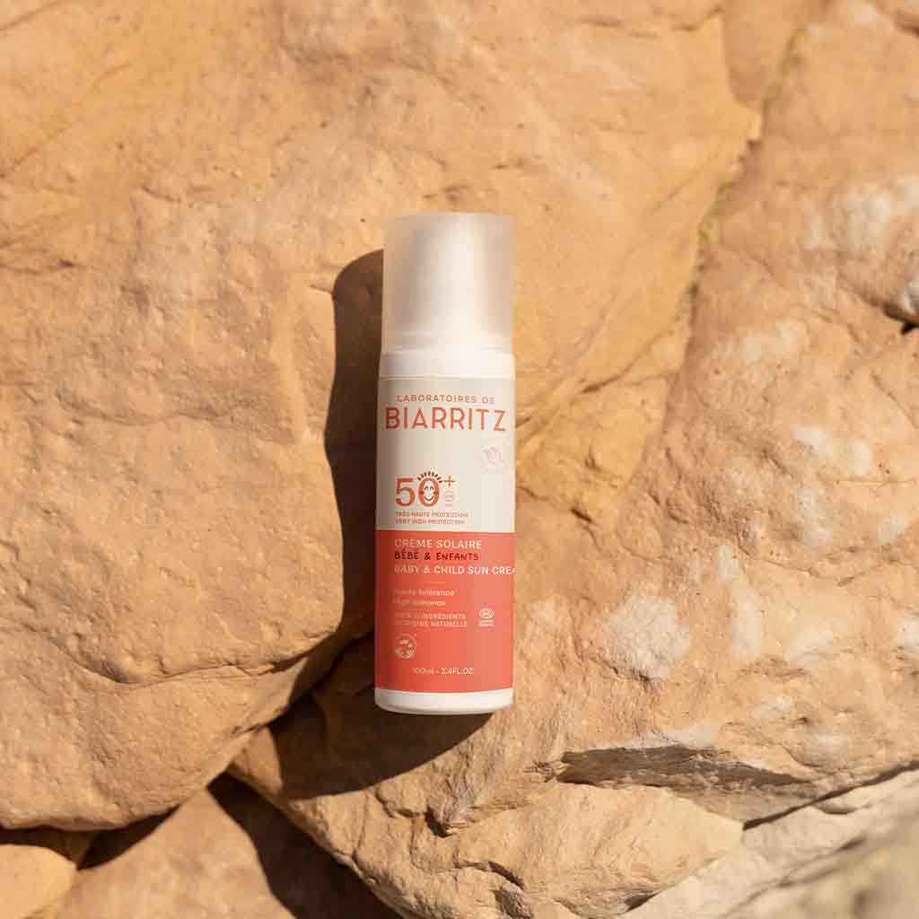 Laboratoires de Biarritz Baby & Child Sun Cream Aurinkovoide lapsille SPF50+; pullo Biarritz SPF 50+ -aurinkovoidetta lepää auringon lämmittämällä, tekstuurisella kivipinnalla ulkona, korostaen laadukasta aurinkosuojaa; pullo valkoinen mattapinnalla ja oranssilla korkilla, sylinterin muotoinen; pakkauksessa lihavoitua groteskitekstiä ”Biarritz” ja ”SPF 50+” selkeästi näkyvissä; ei ihmisiä tai käsiä; luonnollinen ulkovalo valaisee kohtauksen lähikuvassa.