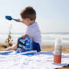 Laboratoires de Biarritz Baby & Child Sun Cream Aurinkovoide lapsille SPF50+; lapsi leikkii hiekalla rannalla istuen valkoisella pyyhkeellä, edustalla pullo SPF 50+ -aurinkovoidetta; muovinen aurinkovoidepullo sylinterin muotoinen kiiltopinnalla, etiketissä ”SPF 50+” lihavoidulla groteskikirjasimella; tuote aseteltu hiekalle pyyhkeen viereen.