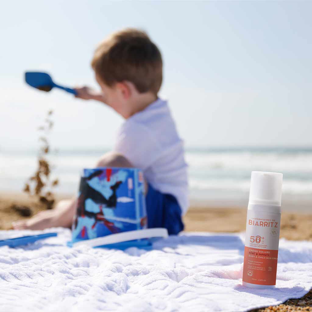 Laboratoires de Biarritz Baby & Child Sun Cream Aurinkovoide lapsille SPF50+; lapsi leikkii hiekalla rannalla istuen valkoisella pyyhkeellä, edustalla pullo SPF 50+ -aurinkovoidetta; muovinen aurinkovoidepullo sylinterin muotoinen kiiltopinnalla, etiketissä ”SPF 50+” lihavoidulla groteskikirjasimella; tuote aseteltu hiekalle pyyhkeen viereen.
