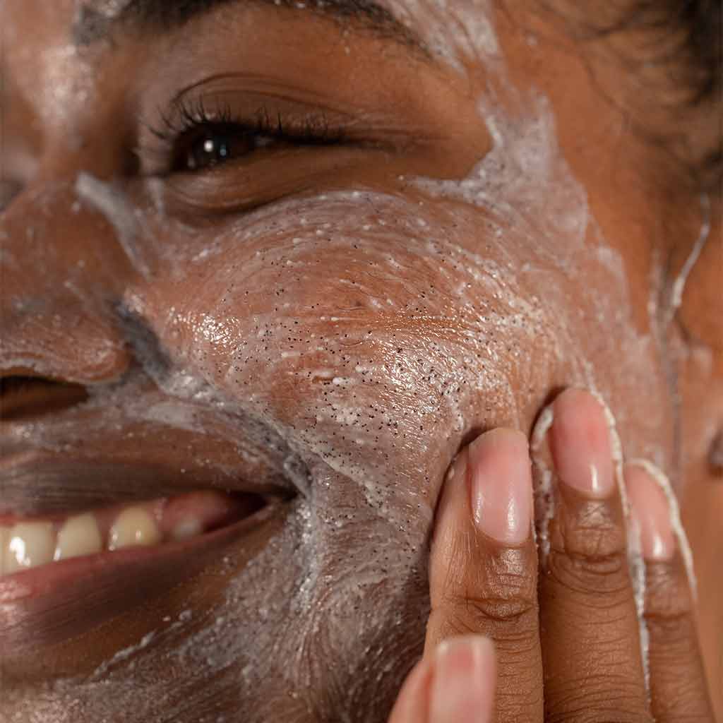 Laboratoires de Biarritz Cleansing Care Kasvokuorinta; henkilö hymyilee levittäessään vaahtoavaa kuorivaa puhdistusainetta hienoin kuorintajyväsin poskelleen sormenpäillä; kirkas geelimäinen puhdistusaine, näkyvissä vaahtoa ja kuorintahiukkasia; ei pakkausta näkyvissä; hyvin valaistu neutraali tausta, tiiviisti rajattu kuva kasvoista ja kädestä; ei bränditekstiä näkyvissä.