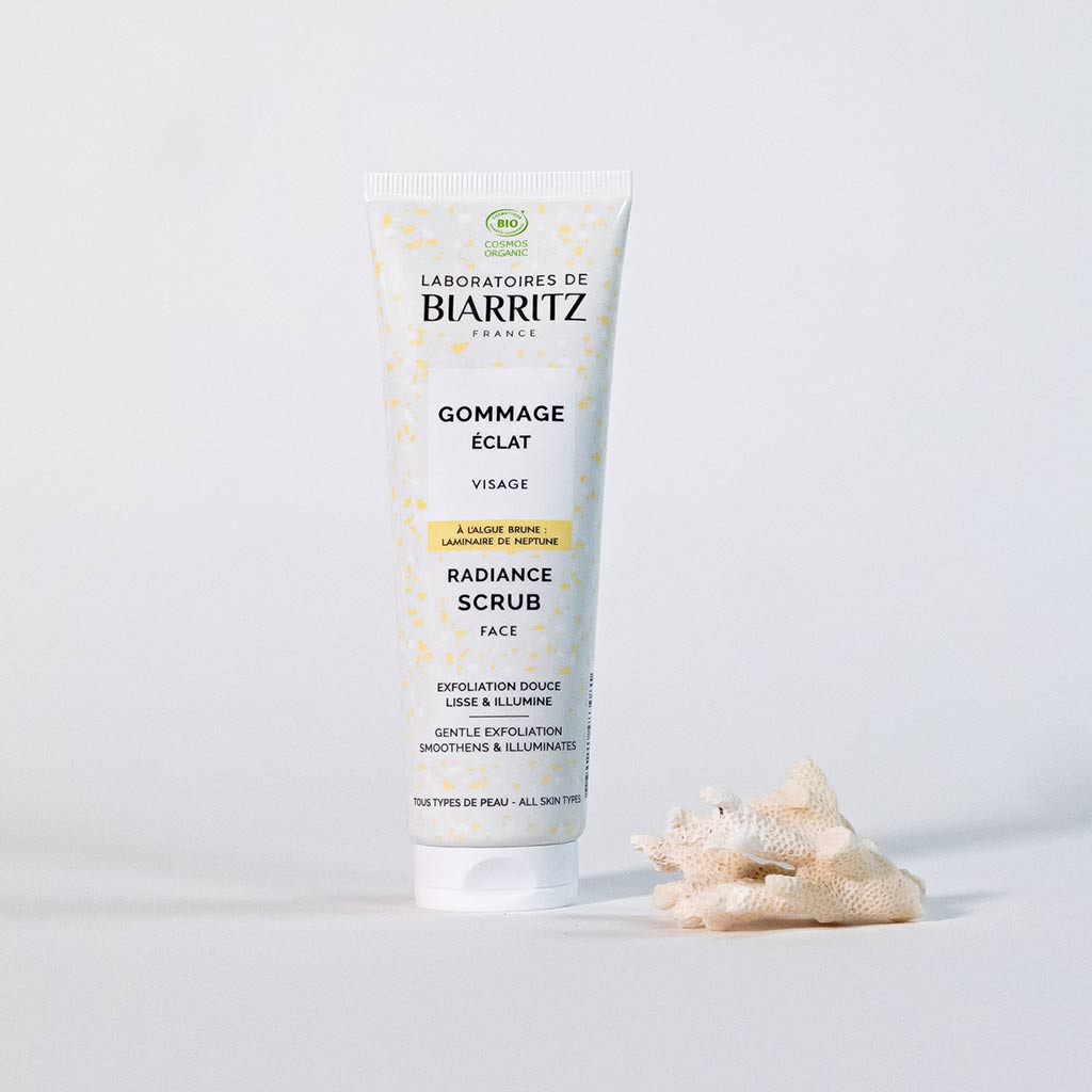 Laboratoires de Biarritz Cleansing Care Kasvokuorinta; tuubi Radiance Scrub -kasvokuorinnalle pystyssä valkoisella alustalla, pieni koralliesine vieressä; tuubi valkoinen mattapintainen turkoosein ja oranssein yksityiskohdin; etiketissä tuotenimi ja brändi groteskifontilla; tausta pysyy tasaisen valkoisena korostaen tätä kuorivaa ihonhoitotuotetta.