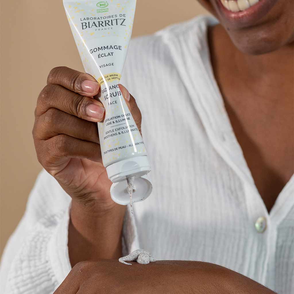 Laboratoires de Biarritz Cleansing Care Kasvokuorinta; henkilö hymyilee purkaessaan kasvokuorintaa pehmeästä valkoisesta muovituubista, jossa luettava teksti "Laboratoires de Biarritz Gommage Eclat", paljaalle käsivarrelleen; pakkaus lieriön muotoinen mattapinnalla ja painetulla groteskitekstillä; henkilöllä yksivärinen valkoinen paita, tausta tasainen beige.