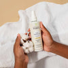 Laboratoires de Biarritz Cleansing Care Puhdistusmaito; kaksi kättä pitelee pulloa Biarritz Cleansing Milk -puhdistusmaitoa ja puuvillankukkaa valkoisen tekstuuripintaisen kangastaustan päällä; pullo lieriön muotoinen, läpinäkymätön valkoinen ja pumppuannostelijalla varustettu; etiketissä brändinimi "Biarritz" groteskifontilla sekä tuotetietoja; kasvot eivät näy; pehmeä luonnonvalo.