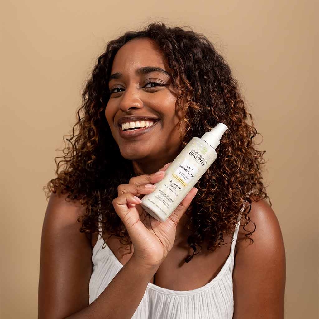 Laboratoires de Biarritz Cleansing Care Puhdistusmaito; hymyilevä kihartuvatukkainen nainen valkoisessa hihattomassa paidassa pitelee lieriön muotoista pulloa tekstillä "Leave-In Conditioner" tasaista beigeä taustaa vasten; pullo pääosin valkoinen kontrastikorkilla ja yksinkertaisella painetulla tekstillä; keskikuva suoraan edestä, ei muita tarvikkeita tai tekstiä.
