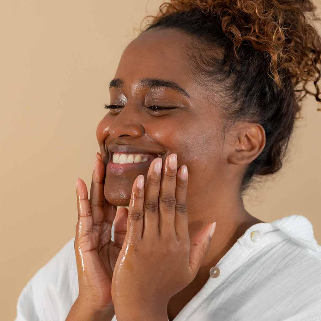 Laboratoires de Biarritz Cleansing Care Puhdistusöljy; kihartuvatukkainen henkilö hymyilee hieroessaan kasvojaan kevyesti ravitsevalla ihonhoitotuotteella; henkilö valkoisessa yläosassa neutraalia taustaa vasten; tuote luonnonvalkoinen voide tai emulsio näkyvissä iholla, ei pakkausta kuvassa; lähikuva päästä ja hartioista, ei tekstiä tai etikettitietoja näkyvissä.
