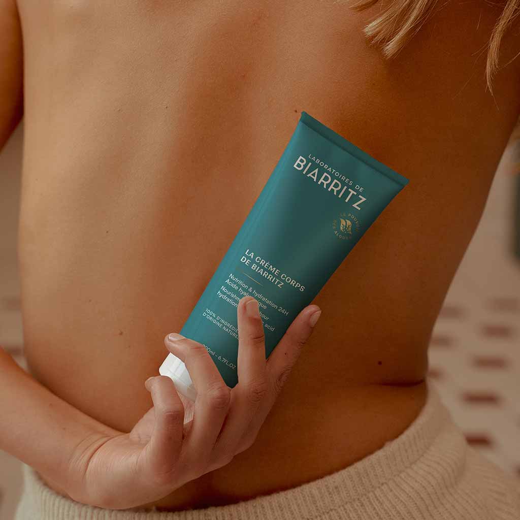 Laboratoires de Biarritz La Crème de Biarritz Body Cream; henkilö paljaalla selällä pitelee sinivihreää Laboratoires de Biarritz -vartalovoidetuubia ihoaan vasten; pukeutuneena beigeihin housuihin, seisoo pehmeästi rajautuvan taustan edessä; tuubi muovia mattapinnalla ja valkoisella korkilla, yksinkertaista mustavalkoista groteskikirjasinta etiketissä; tuotenimi ”Laboratoires de Biarritz” selkeästi näkyvissä tuubin etupuolella.