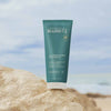 Laboratoires de Biarritz La Crème de Biarritz Body Cream; sinivihreä muovituubi vartalovoidetta mattapinnalla pystyssä karkealla harmaalla kivellä; valkoinen korkki tyvessä; ”Laboratoires de Biarritz” ja tuotetiedot painettu valkoisella groteskikirjasimella etuetikettiin; taustalla epätarkka hiekkaranta ja sininen meri pilvettömän taivaan alla, ei ihmisiä näkyvissä; keskikokoinen lähikuva suoraan edestä.