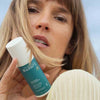 Laboratoires de Biarritz La Crème de Biarritz Face Cream: vaaleahiuksinen nainen pitelee kasvojensa lähellä sylinterimäistä valkoista pulloa, jossa on pumppuannostelija ja mattapintainen etiketti; etiketissä tumma groteskiteksti "Laboratoires Biarritz" ja sinisiä korostuksia; nainen katsoo kameraan; tausta on kirkas, vaaleansininen taivas.