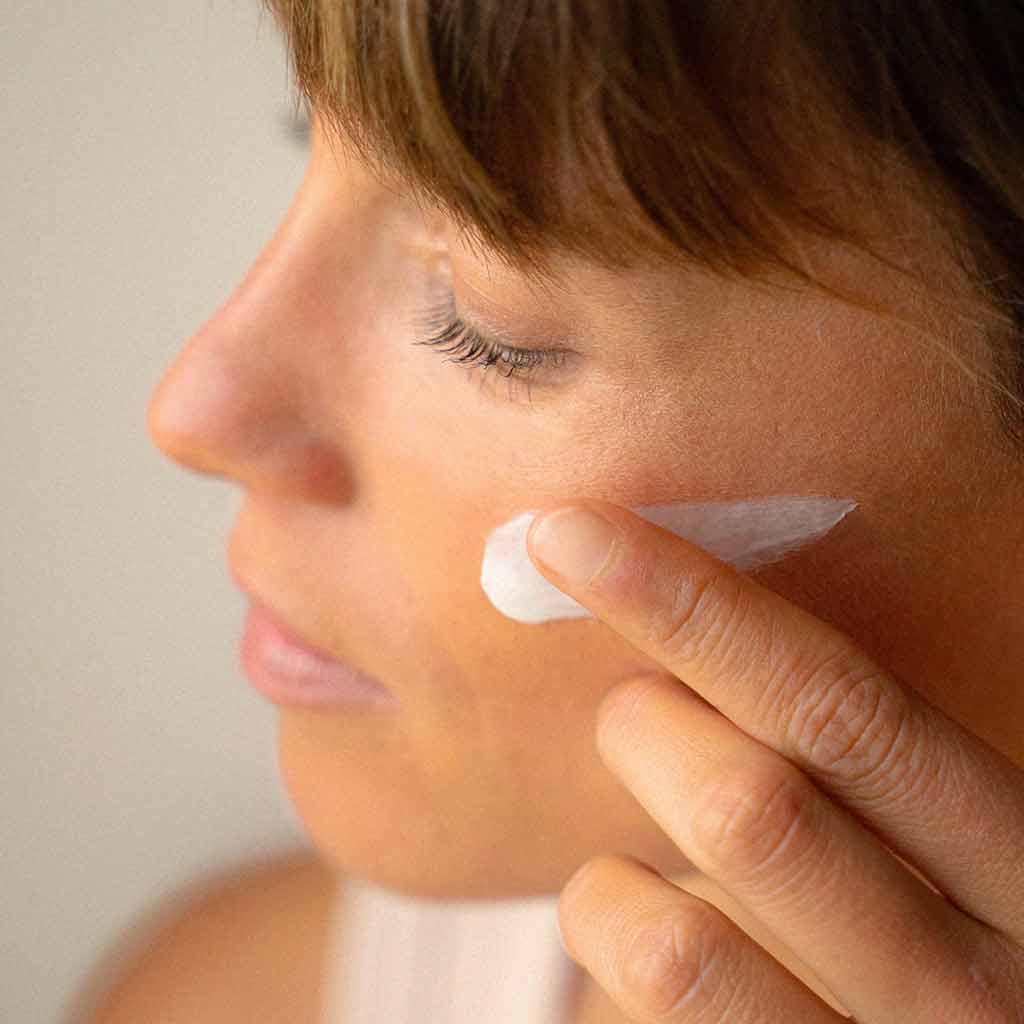 Laboratoires de Biarritz La Crème de Biarritz Face Cream: lähikuva sivuprofiilista, jossa sormella levitetään valkoista, kermaista kosteusvoidetta poskelle; voiteen tekstuuri on läpinäkymätön, kirkkaan valkoinen ja hieman kiiltävä; pakkausta tai etikettejä ei näy; tausta on epätarkka ja neutraali, kuva korostaa kasvoja ja käden liikettä.