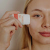 Laboratoires de Biarritz Stick Solaire Aurinkovoidepuikko Valkoinen SPF50+; henkilö pitelee Biarritz SPF 50+ -aurinkovoidepuikkoa lähellä kasvojaan valmistautuen levittämään suojaavaa aurinkosuojatuotetta; kuva keskittyy kasvoihin ja puikkoon; puikko sylinterin muotoinen valkoisessa ja oranssissa muovipakkauksessa mattapinnalla, etiketissä ”Biarritz” lihavoidulla groteskikirjasimella ja ”SPF 50+” selkeästi painettuna; tausta neutraali ja epätarkka; lähikuva.