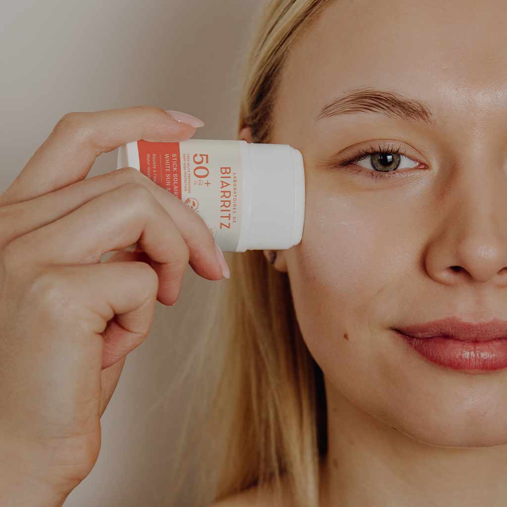 Laboratoires de Biarritz Stick Solaire Aurinkovoidepuikko Valkoinen SPF50+; henkilö pitelee Biarritz SPF 50+ -aurinkovoidepuikkoa lähellä kasvojaan valmistautuen levittämään suojaavaa aurinkosuojatuotetta; kuva keskittyy kasvoihin ja puikkoon; puikko sylinterin muotoinen valkoisessa ja oranssissa muovipakkauksessa mattapinnalla, etiketissä ”Biarritz” lihavoidulla groteskikirjasimella ja ”SPF 50+” selkeästi painettuna; tausta neutraali ja epätarkka; lähikuva.