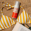 Laboratoires de Biarritz Sun Lotion Aurinkosuojaemulsiovoide SPF30; keltavalkoraidallinen bikiniyläosa aseteltuna tasaisesti hienolle hiekalle SPF 30 -aurinkovoidepullon vierellä, jonka valkoisessa ja oranssissa etiketissä palkkikirjasinta; avoin taskukirja näkyvin tekstiriviein etukansi ylöspäin; ei ihmisiä tai käsiä kuvassa; asetelma ulkona hiekkarannalla ilman häiritseviä taustaelementtejä.