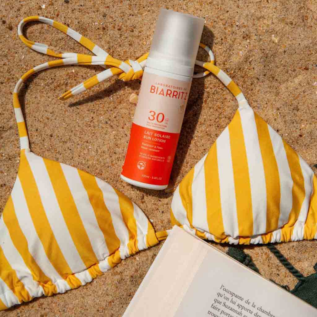 Laboratoires de Biarritz Sun Lotion Aurinkosuojaemulsiovoide SPF30; keltavalkoraidallinen bikiniyläosa aseteltuna tasaisesti hienolle hiekalle SPF 30 -aurinkovoidepullon vierellä, jonka valkoisessa ja oranssissa etiketissä palkkikirjasinta; avoin taskukirja näkyvin tekstiriviein etukansi ylöspäin; ei ihmisiä tai käsiä kuvassa; asetelma ulkona hiekkarannalla ilman häiritseviä taustaelementtejä.
