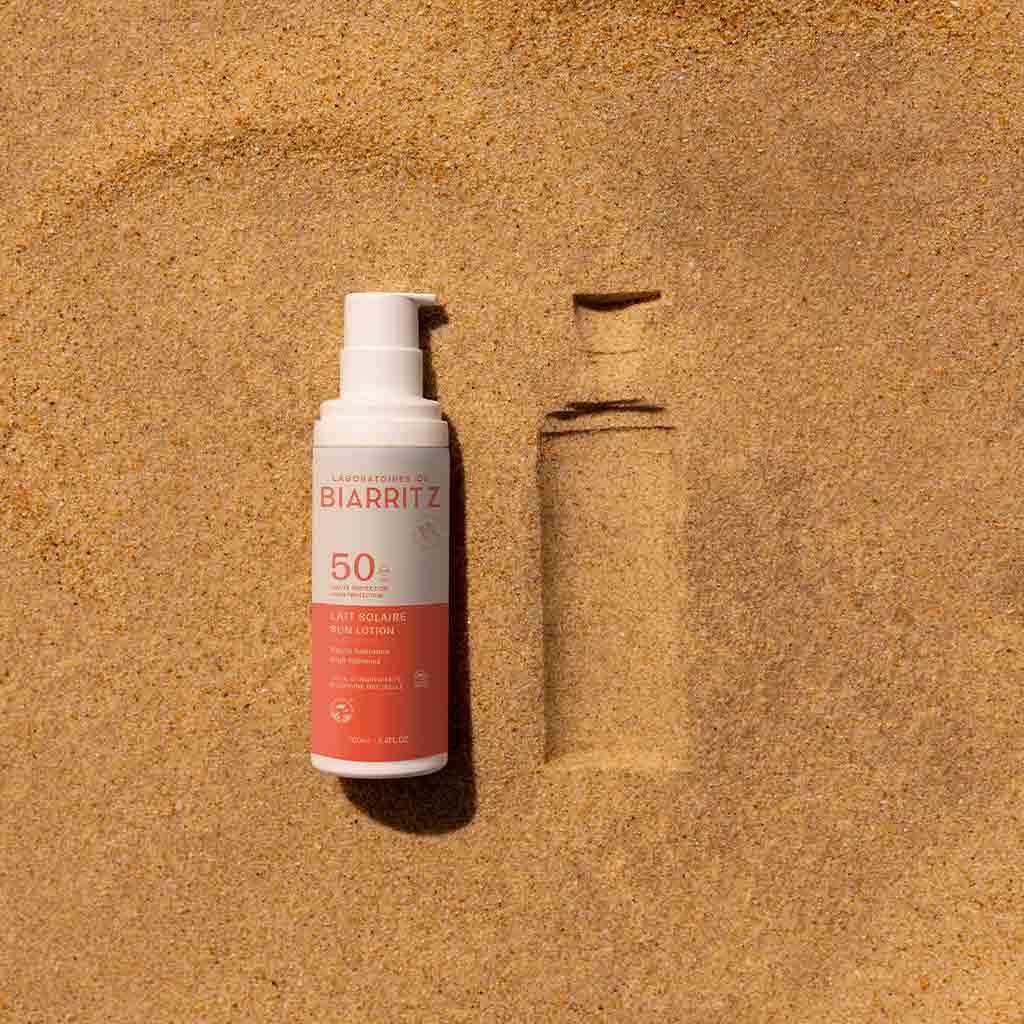 Laboratoires de Biarritz Sun Lotion Aurinkosuojaemulsiovoide SPF50; pullo Biarritz SPF 50 -aurinkovoidetta, sylinterin muotoinen valkoisin ja oranssein sävyin sekä pumppuannostelijalla, seisoo pystyssä hienojakoisella hiekalla sen muotoa vastaavan painauman vieressä; pullo mattamuovipakkauksessa groteskikirjasimin varustetulla etiketillä; ei ihmisiä tai käsiä tässä lähikuvassa; asetelma vihjaa tuotteen juuri sinne asetetuksi.