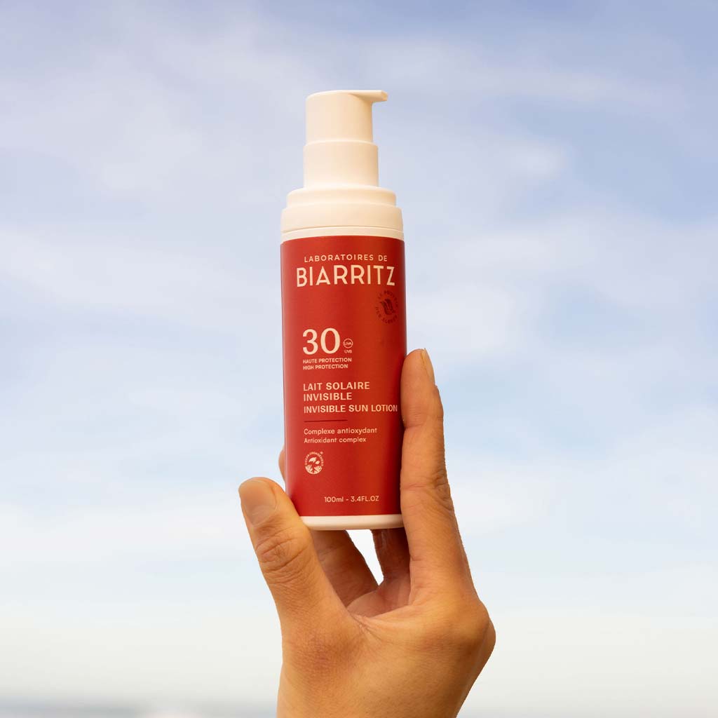 Laboratoires de Biarritz Suncare Invisible Lotion SPF30; käsi pitelee punaista sylinterin muotoista muovipulloa tekstillä ”Laboratoires de Biarritz SPF 30 invisible sun lotion”; pullo mattapintainen valkoisella korkilla ja yksinkertaisella valkoisella groteskitekstillä etupuolella; tausta kirkas sininen taivas; kuvassa näkyy vain henkilön käsi.