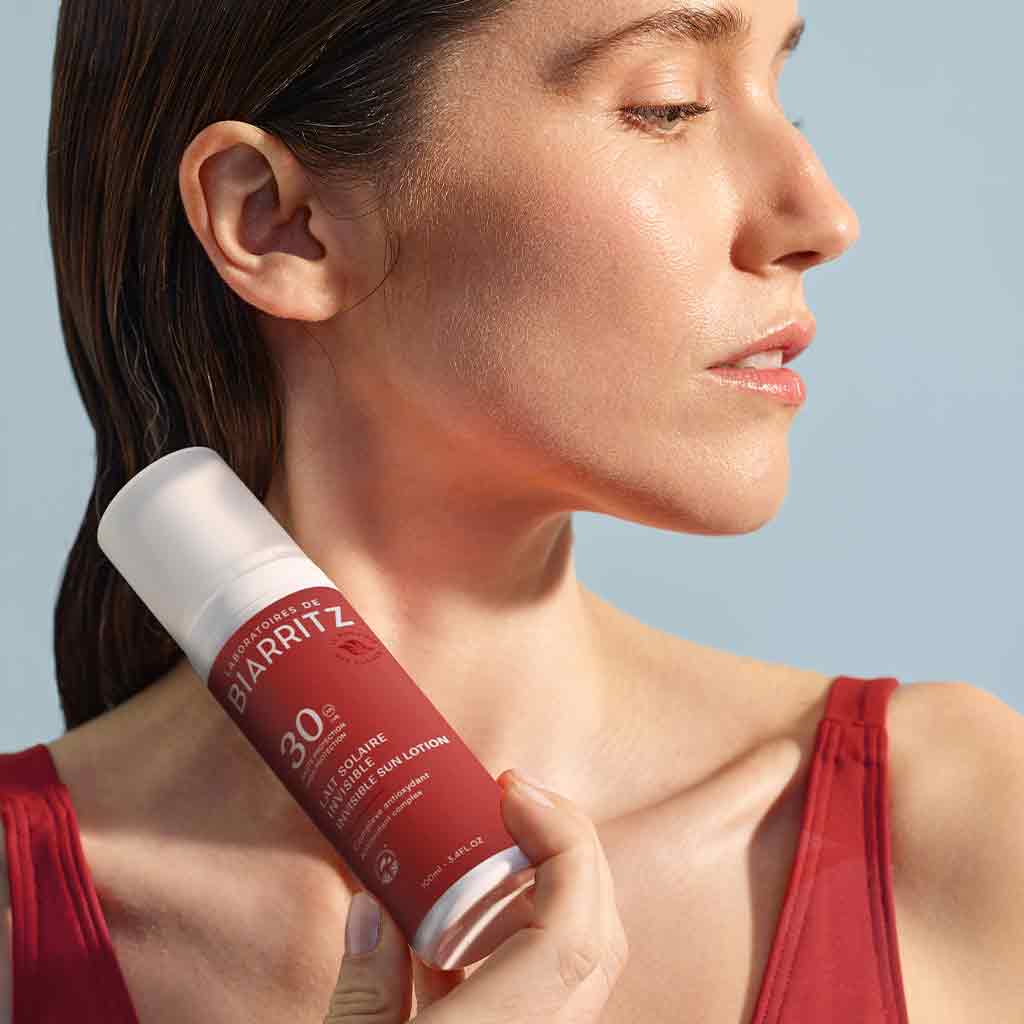 Laboratoires de Biarritz Suncare Invisible Lotion SPF30; henkilö punaisessa yläosassa pitelee sylinterin muotoista punaista muovipulloa lähellä kaulaansa; etiketissä valkoinen teksti ”Biarritz SPF 30” selkeästi näkyvissä; pullon pinta matta ja korkki valkoinen; tausta tasaisesti valaistu vaaleansininen; henkilö katsoo kuva-alan ulkopuolelle, vain yläkroppa näkyvissä kuvassa.