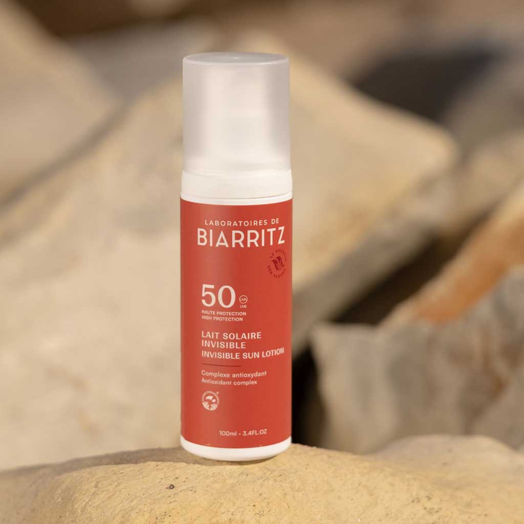 Laboratoires de Biarritz Suncare Invisible Lotion SPF50; valkoinen sylinterin muotoinen pullo Laboratoires de Biarritz SPF 50 -näkymätöntä aurinkovoidetta mattapinnalla seisoo pystyssä tasaisella beigellä kivellä; korkki myös valkoinen ja vaikuttaa muovilta; etikettiteksti näyttää brändinimen ja SPF-tiedot yksinkertaisella groteskikirjasimella; sumeana taustalla tekstuurisia kiviä sinivihrein mineraalikerrostumin; ei ihmisiä tai käsiä.