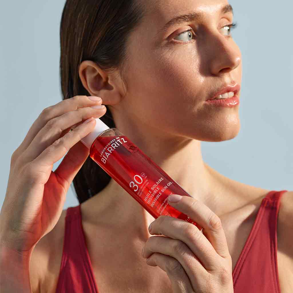 Laboratoires de Biarritz Suncare Invisible Oil SPF30; henkilö punaisessa yläosassa pitelee sylinterin muotoista punaista pulloa tekstillä Biarritz SPF 30 aurinkosuoja, valkoista typografiaa pullossa; pakkauksen pinta matta; kuva vyötäröstä ylöspäin, henkilö katsoo sivulle; tausta yhtenäisen vaaleansininen ilman muita esineitä.