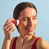 Laboratoires de Biarritz Suncare Invisible Sun Stick SPF50+; henkilö punaisessa yläosassa hymyilee levittäessään aurinkosuojapuikkoa kasvoilleen vaaleansinistä taustaa vasten; aurinkosuoja sylinterin muotoisena kiinteänä puikkona muovipakkauksessa, mattavalkoinen korkki ja värillinen pohja; yksinkertaista groteskitekstiä näkyvissä etiketissä; ei muita esineitä tai rekvisiittaa; kuva vyötäröstä ylöspäin, tarkennus levitystoiminnossa.
