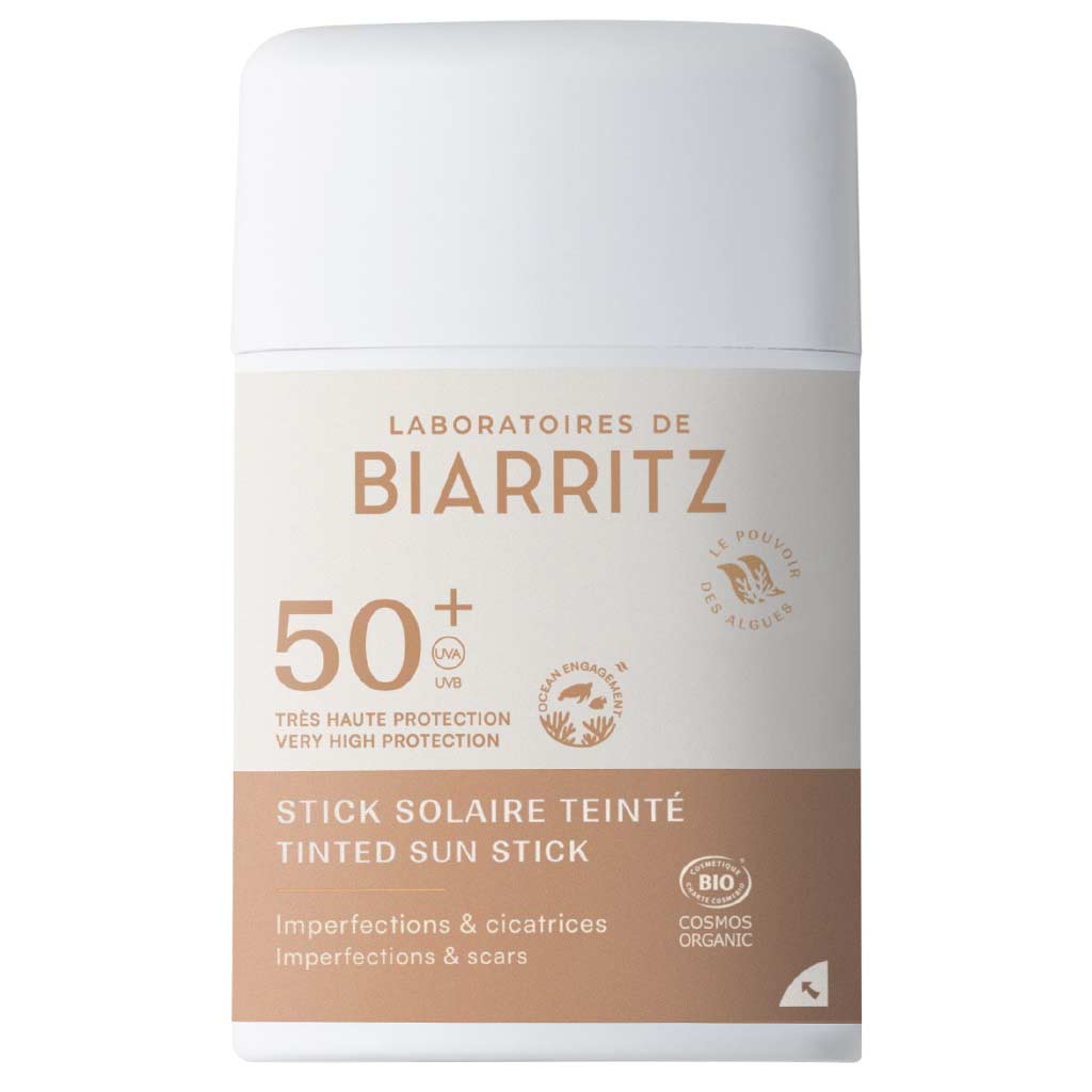 Laboratoires de Biarritz Tinted Sunscreen Stick SPF50+ (Sävytetty Aurinkovoidepuikko): valkoinen ja beige sylinterimäinen puikko mattapinnalla, lihavoitu groteskiteksti; etiketissä maininnat epäpuhtauksien ja arpien peittämisestä, luomusertifikaattien logot sekä sävytetty suoja; muovinen pakkaus, tuotetiedot painettu suoraan etikettiin; tausta on valkoinen, tuote keskellä kuvaa, ei ihmisiä tai käsiä näkyvissä.