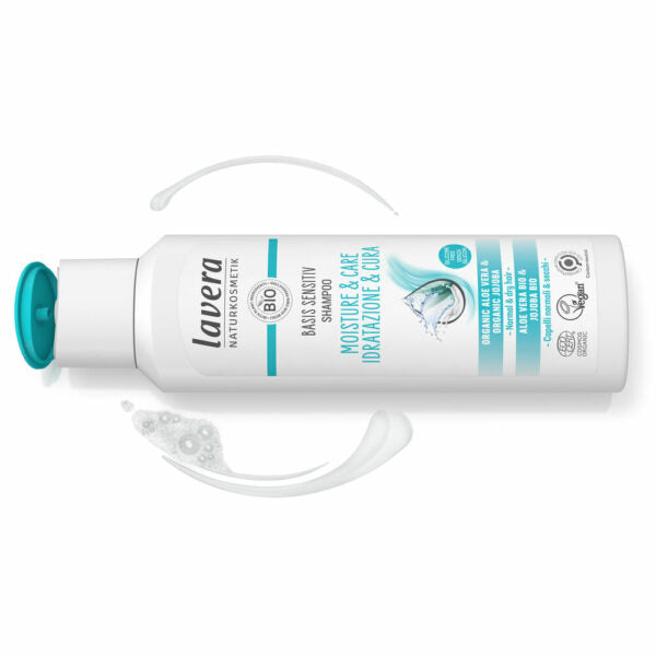 Valkoinen Lavera Moisture & Care -shampoopullo avoimella turkoosilla läppäkorkilla, vieressä tuotenäyte, sinisin etikettitekstein kuvattuna sen hellävaraisia ja kosteuttavia ominaisuuksia herkälle iholle; pullo mattapintainen ja lieriömäinen; sininen groteskikirjasin etiketissä; ei ihmisiä tai käsiä näkyvissä; tuote kuvattu vaalealla taustalla läheltä.