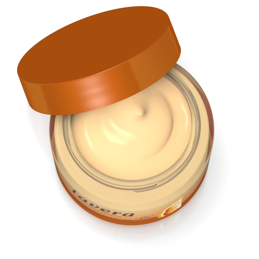 Lavera Glow By Nature Vitamin C 3in1 Cream; avattu voidepurkki oranssilla muovikannella ja pyöreällä sileäsivuisella rungolla; purkissa beigeä voidetta kiiltävällä pinnalla; osittain näkyvä etiketti käyttää yksinkertaista groteskikirjasinta mustalla tekstillä valkoista taustaa vasten, luettava teksti vähäistä; tuote valkoisella taustalla, keskikokoinen lähikuva, ei ihmisiä tai käsiä.