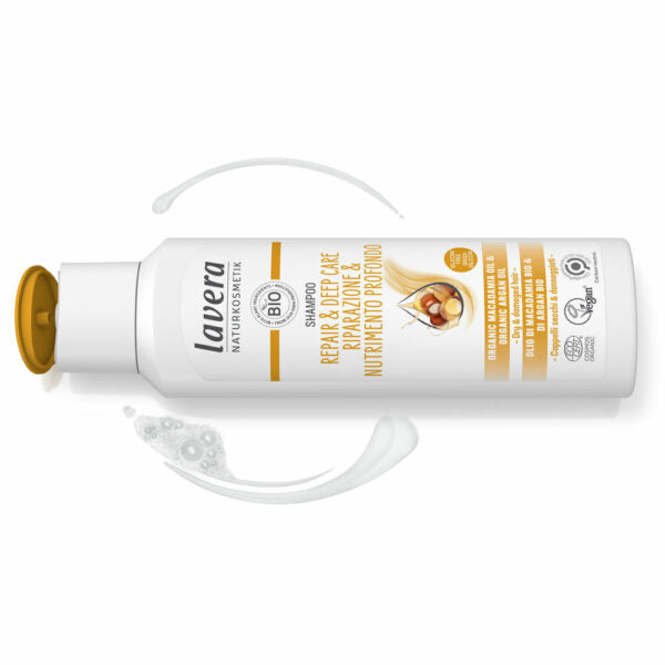 Valkoinen Lavera Repair & Deep Care Shampoo -pullo avoimella keltaisella korkilla; pinta sileä ja mattainen, kulmat pyöristetyt; etiketissä mustaa ja kultaista tekstiä argaanöljystä ja luomusertifikaateista; pullo on aseteltu valkoiselle taustalle kahden kermaisen shampoolevitteen viereen; ei ihmisiä tai käsiä näkyvissä kuvassa.