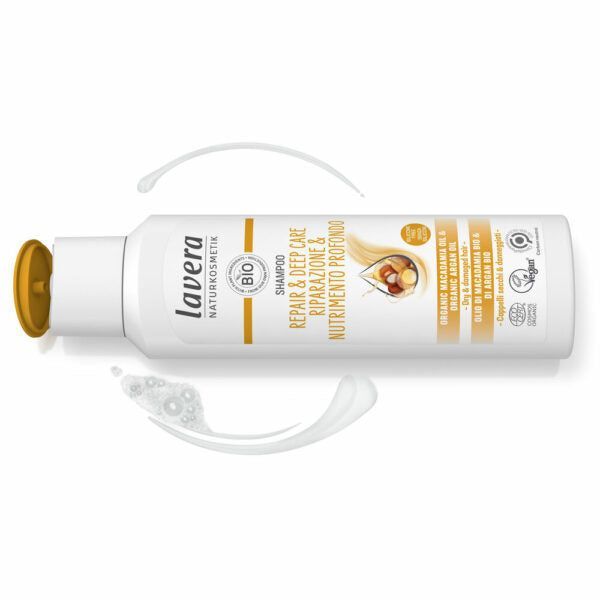 Valkoinen Lavera Repair & Deep Care Shampoo -pullo avoimella keltaisella korkilla; pinta sileä ja mattainen, kulmat pyöristetyt; etiketissä mustaa ja kultaista tekstiä argaanöljystä ja luomusertifikaateista; pullo on aseteltu valkoiselle taustalle kahden kermaisen shampoolevitteen viereen; ei ihmisiä tai käsiä näkyvissä kuvassa.