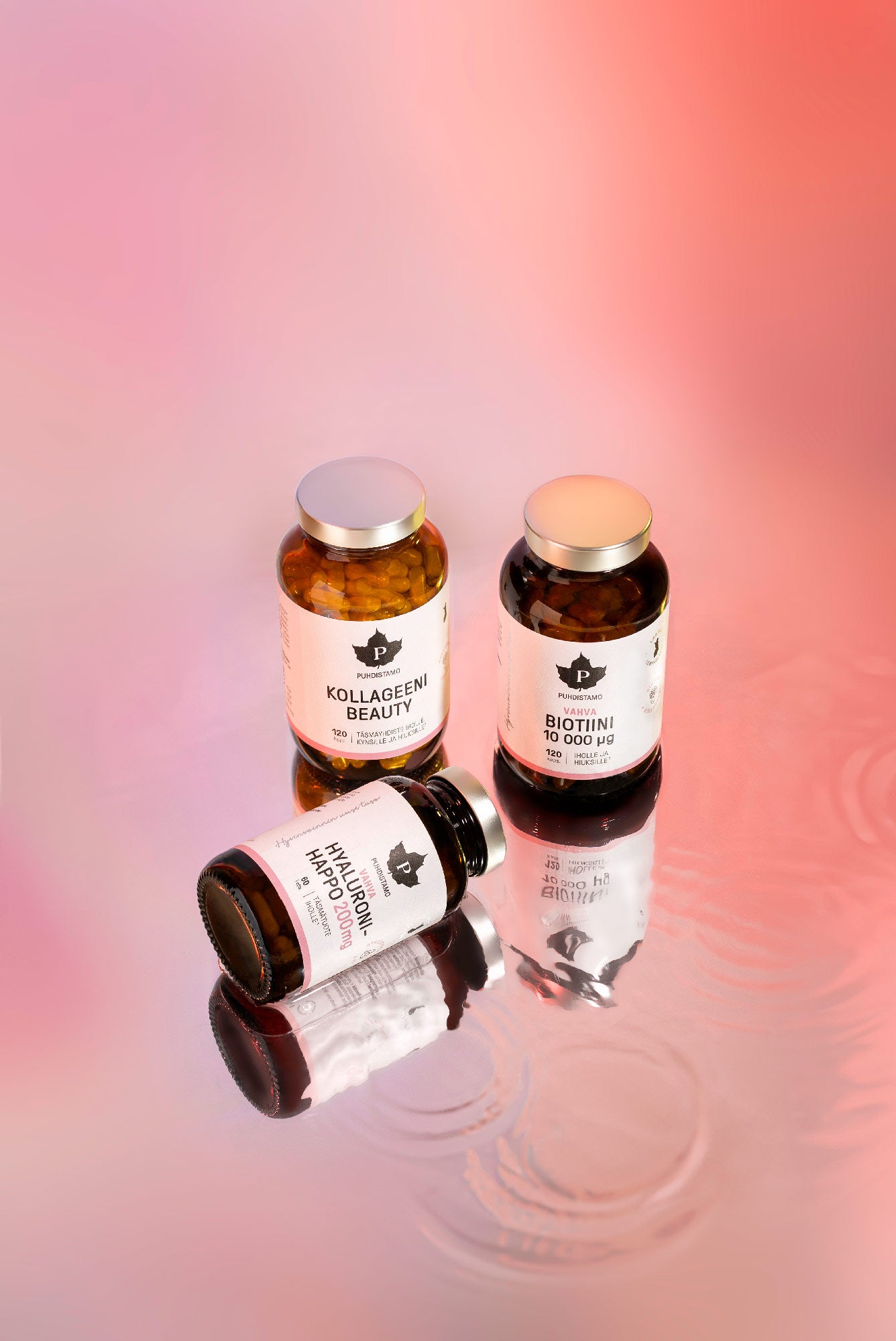 Puhdistamo Beauty Box; kolme ravintolisäpulloa nimillä BIOTIINI, KOLLAGEENI ja MULTIVITAMIINI aseteltuna vaaleanpunais-persikansävyiselle liukuvärialustalle, yksi pullo vaakatasossa ja kaksi pystyssä; kaikki pullot lieriömäisiä valkoista mattapintaista muovipakkausta; etiketeissä lihavoitua groteskityypografiaa; pintaan heijastuu lievästi pullojen kuva; ei ihmisiä tai käsiä kuvassa.