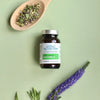 Living Nutrition Fermentoitu Maarianohdake; sylinterinmuotoinen muovipullo merkitty "Living Nutrition Milk Thistle", valko-vihreä väritys ja groteskikirjasin, seisoo pystyssä mattavihreällä pinnalla; vieressä puulusikka täynnä irtonaisia kuivattuja yrttejä ja eloisan violetti kukkatertti; ei ihmisiä tai käsiä näkyvissä; pullo lepää vihreällä alustalla puulusikan ja kukkien vieressä korostaen luonnollisia ainesosia.