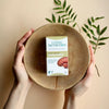 Living Nutrition Fermentoitu Reishi; kädet pitelevät pyöreää, mattapintaista puukulhoa, jossa suorakaiteen muotoinen rasia tekstillä ”Living Nutrition Organic Fermented Reishi Spores” selkeällä groteskikirjasimella; rasia valkoinen ja oranssi, näyttää tuotenimen ja brändin; kaksi tuoretta vihreää lehteä lepää kulhon vieressä vaalealla, beigellä alustalla; kuva ylhäältä, ei muita esineitä näkyvissä.