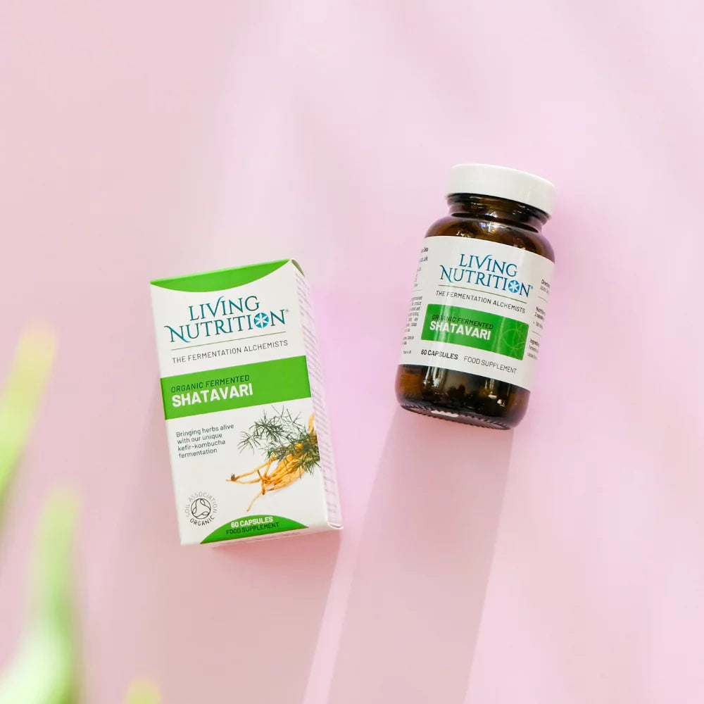 Living Nutrition Fermentoitu Shatavari; laatikko ja pullo Living Nutrition -luomufermentoituja Shatavari-kapseleita on aseteltu vaaleanpunaiselle pinnalle, muutamia vihreitä lehtiä kulmassa; laatikko on suorakulmainen mattapinnalla, pääosin valkoinen turkoosin ja tummansinisin korostein groteskikirjasimella; pullo on ruskeaa lasia valkoisella etiketillä samaa brändäystä; molemmat esineet avaamattomia ja pystyssä; ei ihmisiä tai käsiä näkyvissä; keskikokoinen kuva ylhäältä.