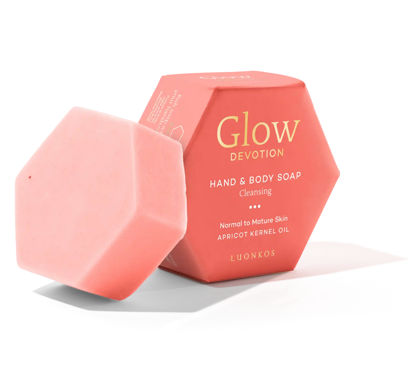 Luonkos Glow Devotion Hand & Body Soap; vaaleanpunainen kuusikulmainen saippuapala nojaa yhteensopivaa kuusikulmaista rasiaansa vasten, jossa teksti ”Glow Devotion Hand & Body Soap: Cleansing, Apricot Kernel Oil, Luonkos”, suunniteltu normaalille ja kypsälle iholle; sekä saippua että rasia mattapintaisia; rasian typografia lihavoitua tikkukirjasinta; kuvausympäristö tasainen valkoinen, ei ihmisiä tai muita esineitä näkyvissä.