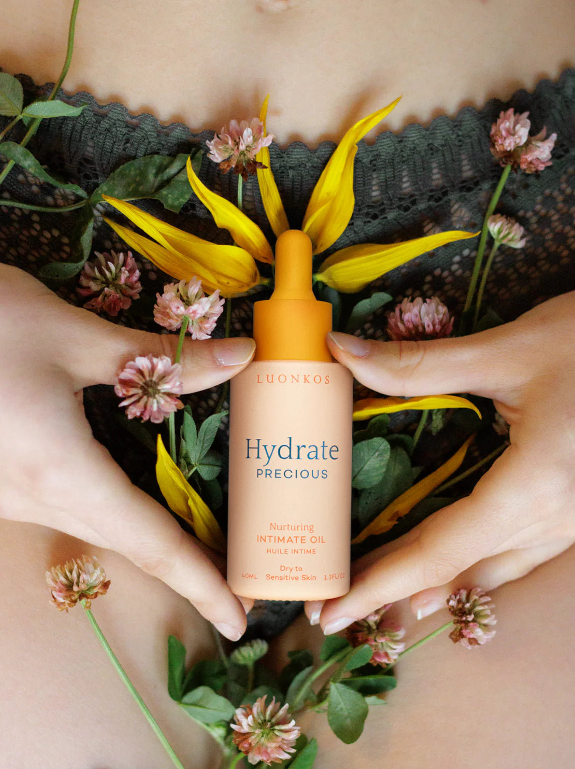 Käsi pitelee pientä lieriömäistä pulloa, jossa lukee Luonkos Hydrate Precious Intimate Oil valkoisella tekstillä ja minimalistisella muotoilulla; pullo mattapintainen, pidetään paljasta ihoa vasten; käden ympärillä keltaisia kukanterälehtiä ja violetteja apilankukkia; kuvan kulmassa näkyy vihreää pitsikangasta; lähikuva.