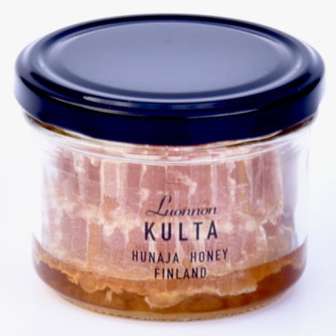 Luonnon Kulta Kennohunaja; lasipurkki tummalla kannella sisältää mehiläiskennoa ja hunajaa; läpinäkyvä purkki on sylinterinmuotoinen sileine sivuineen ja kiiltäväpintainen; edessä oleva etiketti sisältää tekstin ”Luonnon Kulta Hunaja Honey Finland”, korostaen puhdasta suomalaista hunajaa; etiketti käyttää selkeitä groteskifontteja mustan ja kullan sävyissä; tuote kuvattu valkoista taustaa vasten hyvin valaistussa studiokuvassa; ei ihmisiä tai käsiä näkyvissä.