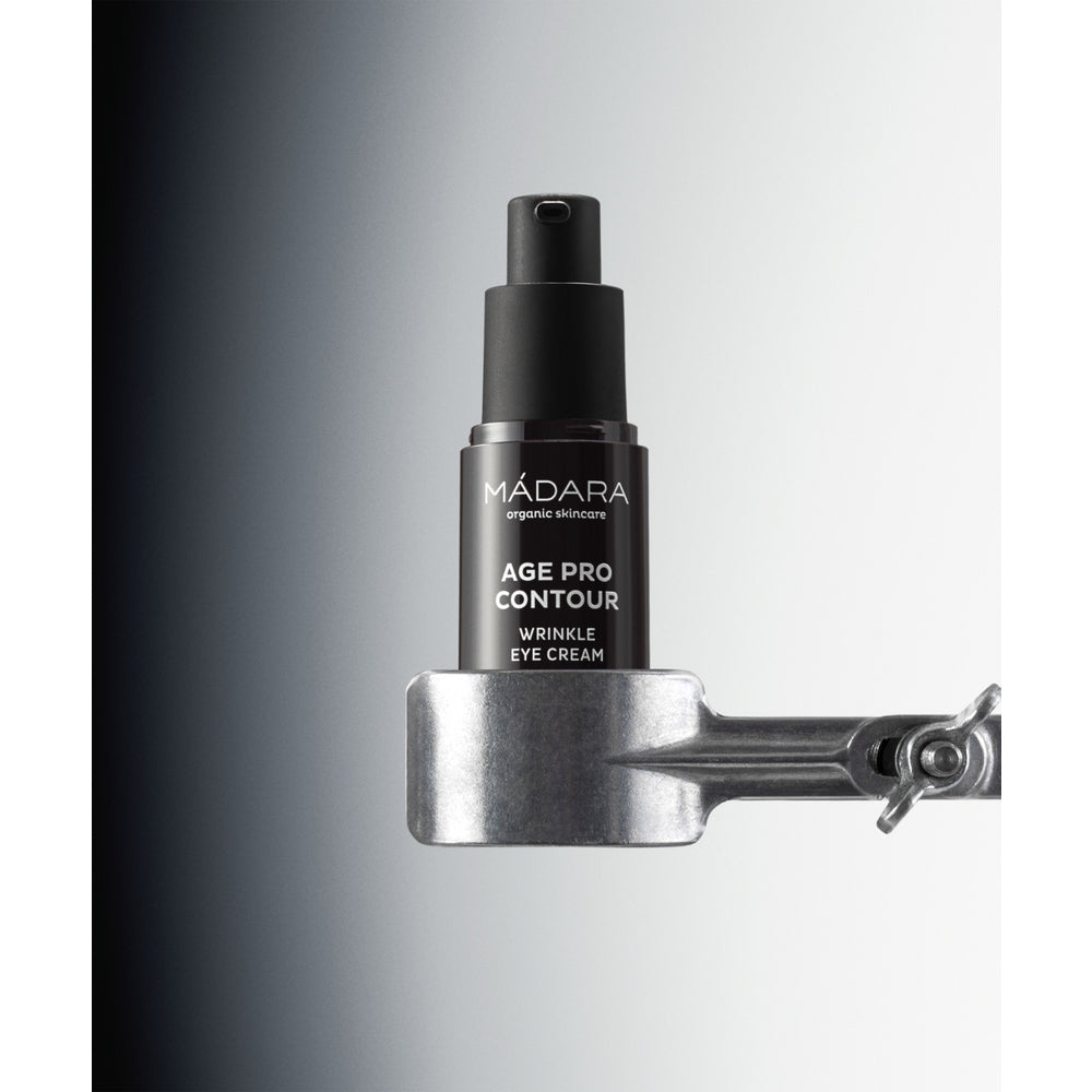 Madara AGE PRO Contour Wrinkle Eye Cream; musta pullo MÁDARA Age Pro Contour Wrinkle Eye Cream -tuotetta on kiinnitetty metalliseen pidikkeeseen liukuväritaustaa vasten, joka häivytetään tummanharmaasta vasemmalta valkoiseen oikealla, korostaen tuotteen tyylikästä muotoilua; pullo on mattapintainen ja sylinterinmuotoinen, etuetiketissä valkoista groteskitekstiä tuotenimestä; ei ihmisiä tai käsiä näkyvissä; studiokuva.
