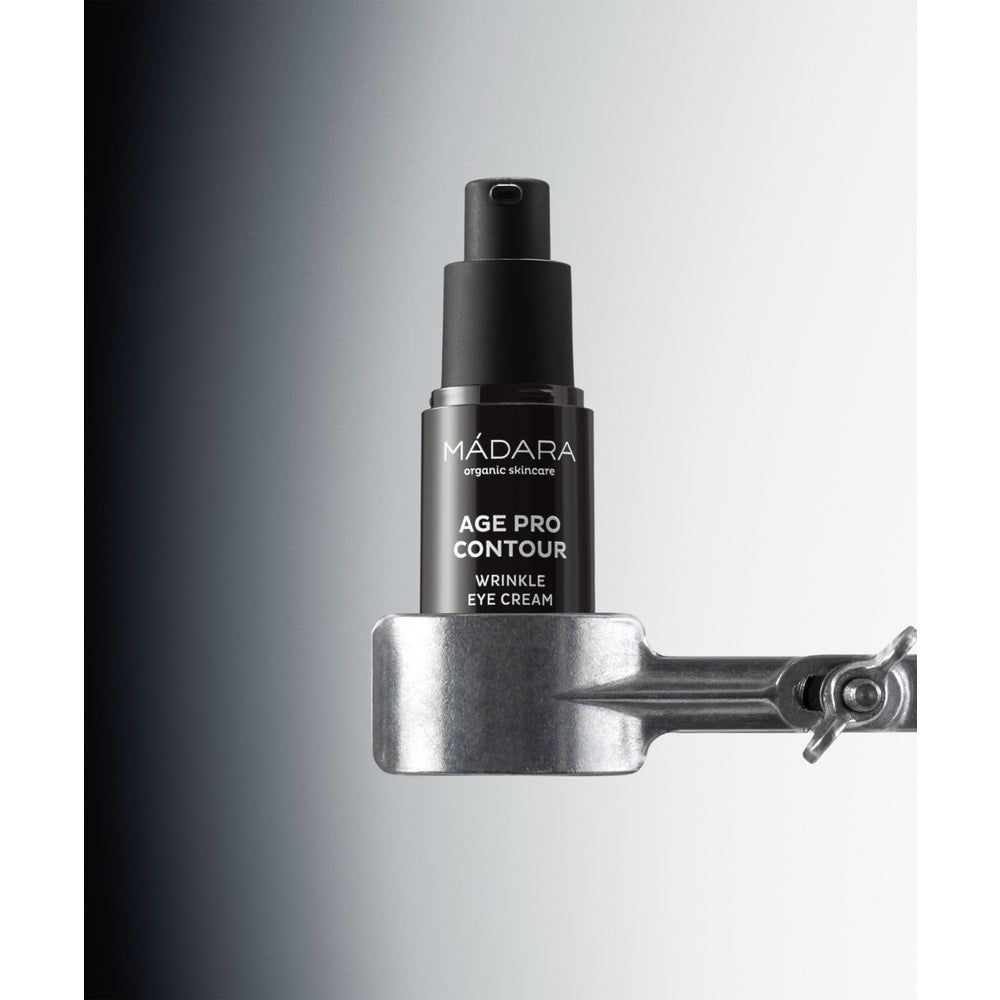 Madara AGE PRO Contour Wrinkle Eye Cream; musta pullo MÁDARA Age Pro Contour Wrinkle Eye Cream -tuotetta on kiinnitetty metalliseen pidikkeeseen liukuväritaustaa vasten, joka häivytetään tummanharmaasta vasemmalta valkoiseen oikealla, korostaen tuotteen tyylikästä muotoilua; pullo on mattapintainen ja sylinterinmuotoinen, etuetiketissä valkoista groteskitekstiä tuotenimestä; ei ihmisiä tai käsiä näkyvissä; studiokuva.