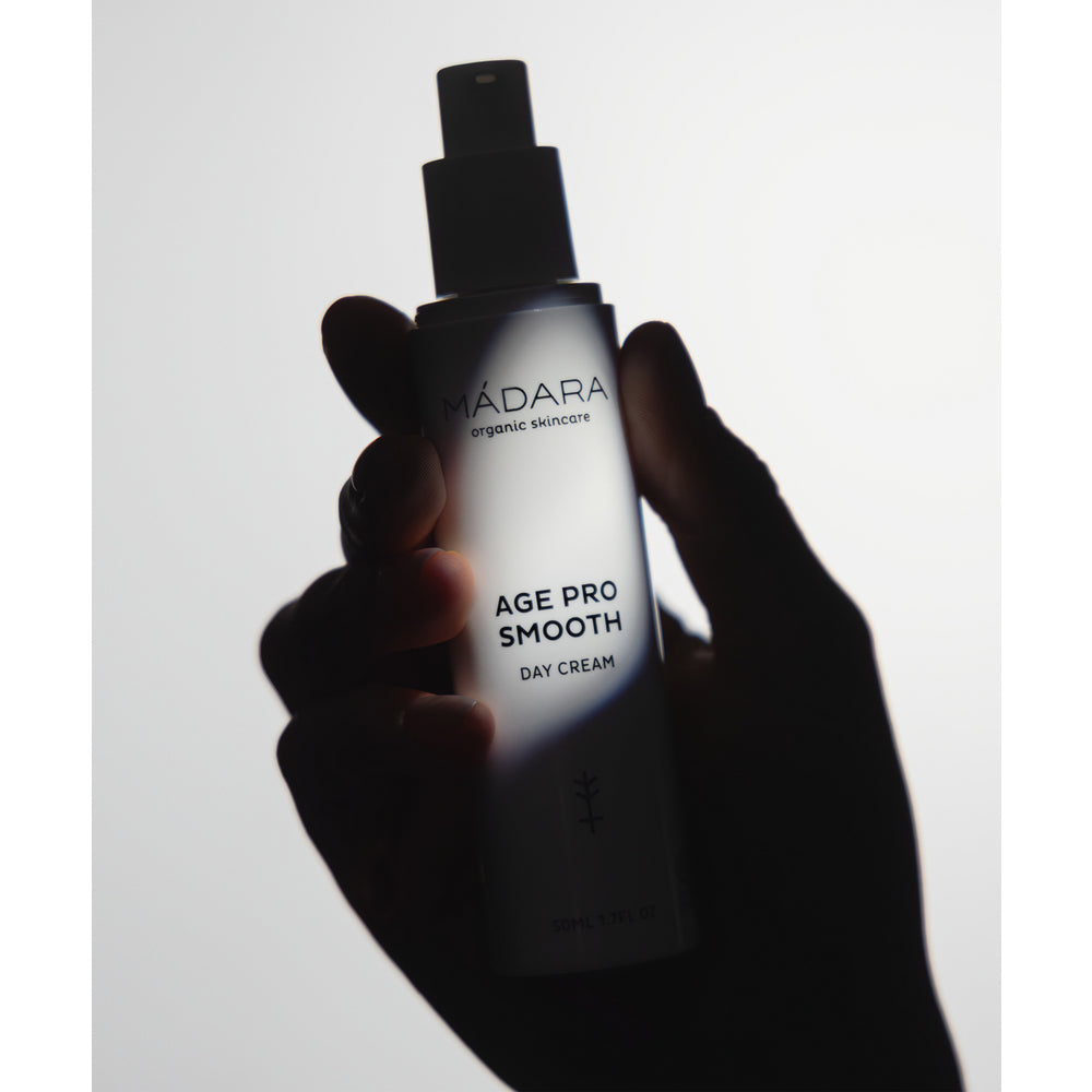 Madara AGE PRO Smooth Day Cream; käsi pitelee Mádara Organic Skincare Age Pro Smooth Day Cream -pulloa valkoista taustaa vasten, luonnonvalo valaisee pehmeästi tätä luonnonkosmetiikkatuotetta; pullo on sylinterinmuotoinen mattavalkoisella pinnalla ja pystysuuntaisella minimalistisella mustalla tekstillä; etiketti on luettavissa: ”Mádara Organic Skincare Age Pro Smooth Day Cream”; ei muita ylimääräisiä elementtejä näkyvissä.