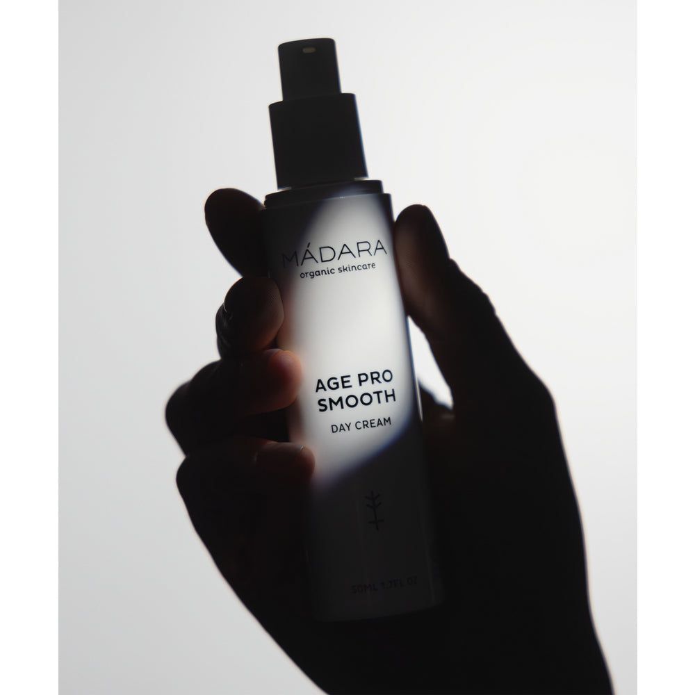 Madara AGE PRO Smooth Day Cream; käsi pitelee Mádara Organic Skincare Age Pro Smooth Day Cream -pulloa valkoista taustaa vasten, luonnonvalo valaisee pehmeästi tätä luonnonkosmetiikkatuotetta; pullo on sylinterinmuotoinen mattavalkoisella pinnalla ja pystysuuntaisella minimalistisella mustalla tekstillä; etiketti on luettavissa: ”Mádara Organic Skincare Age Pro Smooth Day Cream”; ei muita ylimääräisiä elementtejä näkyvissä.
