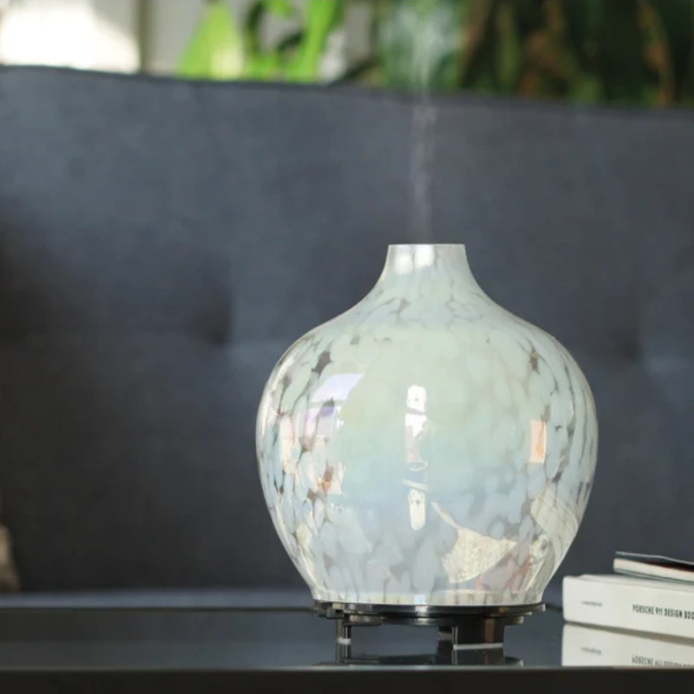Made by Zen Mercura Diffuuseri; keraaminen aromidiffuusori seisoo pöydällä tumman, nypytetyn sohvan edessä, lähellä pinottuja kirjoja ja viherkasveja taustalla; höyryä nousee kevyesti diffuusorin yläosasta; diffuusori on mattapintainen ja pyöreähkö; ei näkyvää pakkausta tai etikettiä; kuva otettu sisätiloissa ilman ihmisiä tai käsiä.