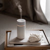 Made by Zen Nomad USB Diffuuseri; valkoinen sumua höyryävä sylinterinmuotoinen diffuusori suorakulmaisella tarjottimella; tarjottimella myös suljettu hopeanvärinen kannettava tietokone, suuri luonnonvärinen simpukankuori ja avattu aikakauslehti tekstein ja kuvin; kaikki aseteltu sileälle pinnalle pehmeästi valaistussa huoneessa beigein seinin ja minimalistisin sisustuksin; ei ihmisiä tai käsiä näkyvissä.
