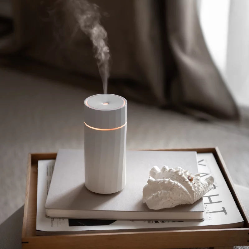 Made by Zen Nomad USB Diffuuseri; valkoinen sumua höyryävä sylinterinmuotoinen diffuusori suorakulmaisella tarjottimella; tarjottimella myös suljettu hopeanvärinen kannettava tietokone, suuri luonnonvärinen simpukankuori ja avattu aikakauslehti tekstein ja kuvin; kaikki aseteltu sileälle pinnalle pehmeästi valaistussa huoneessa beigein seinin ja minimalistisin sisustuksin; ei ihmisiä tai käsiä näkyvissä.