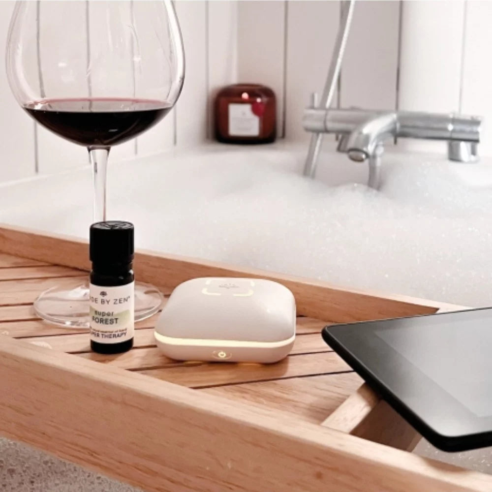 Made by Zen Peri USB Diffuuseri; lasi punaviiniä, pieni eteerisen öljyn pullo, diffuusori ja tabletti lepäävät luonnollisen syyllisen puisella kylpyammetarjottimella kylpyammeen päällä täynnä valkoista vaahtoa; eteerisen öljyn pullo on kirkasta lasia tummalla kierrekorkilla ilman luettavaa etikettiä; diffuusori on sylinterinmuotoinen ja mattavalkoinen; taustalla kromisen hanan lähellä palaa merkitsemätön kynttilä pehmeästi; kuvassa ei ole ihmisiä tai käsiä.