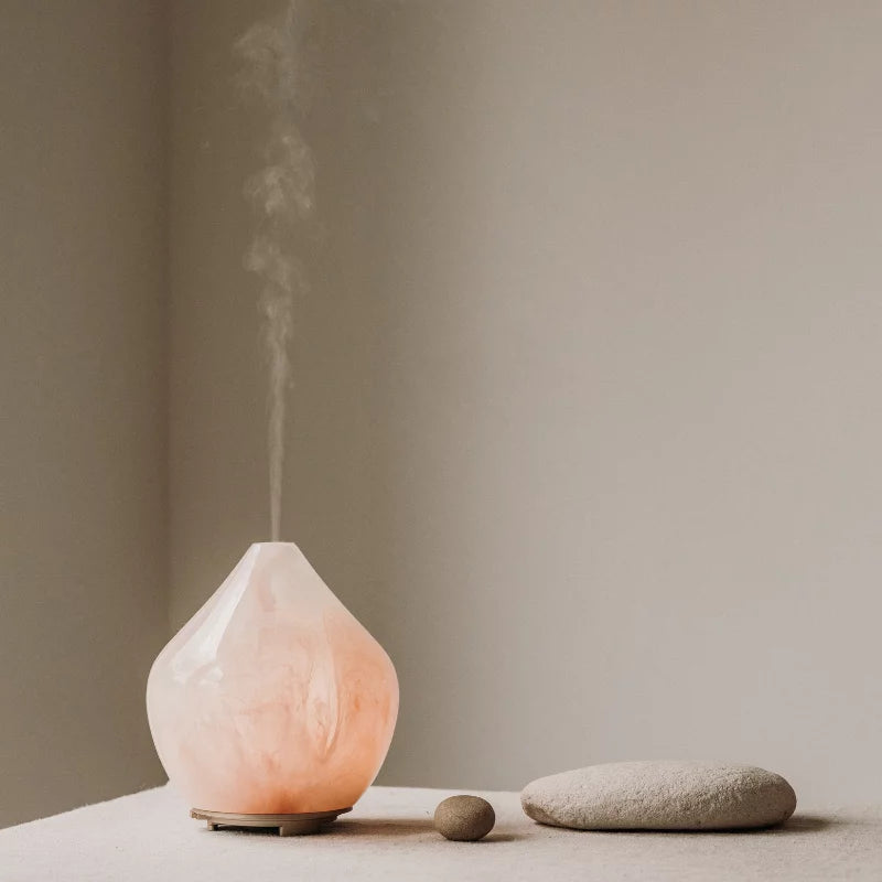 Made by Zen Gem Diffuuseri; vaaleanpunainen marmorikuvioinen diffuusori höyryää ohuen sumuvirtauksen, aseteltuna vaalean kangaspinnan päälle kahden sileän beigen kiven viereen neutraalia taustaa vasten; diffuusori on pyöreä ja mattapintainen, jossa näkyy pehmeitä vaaleanpunaisia ja valkoisia marmorikuvioita; ei näkyvää etikettiä tai luettavaa tekstiä; asetelma on minimalistinen ilman ihmisiä tai käsiä, kuvattu suoraan edestä silmän korkeudelta.