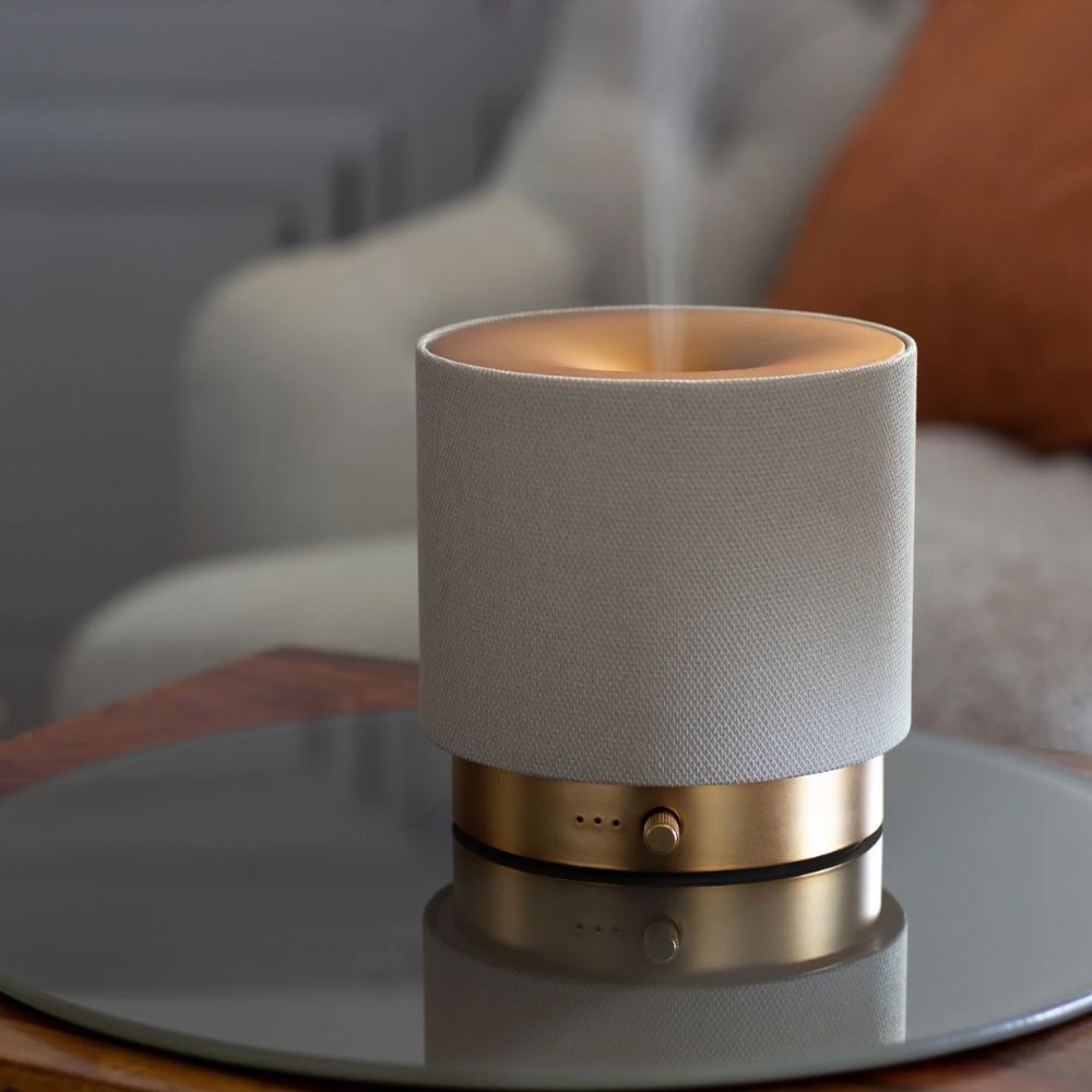 Made by Zen Mira USB Diffuuseri; moderni, lieriömäinen aromidiffuusori kultaisella metallijalustalla ja beigellä kangaspintaisella suojuksella, päästää kapean höyryvirran; pinta mattapintainen kankaassa ja heijastava kultajalustassa; ei näkyviä etikettejä tai tekstiä; tuote lepää kiiltävällä, peilimäisellä pöytäpinnalla sisustetussa sisätilassa; ei ihmisiä kuvassa.