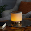 Made by Zen Mira USB Diffuuseri; lieriömäinen lamppu pehmeine lämpimine hehkuineen seisoo pyöreällä peilillä puupinnan päällä; lampun runko läpinäkymätön ilman näkyvää brändäystä tai merkintöjä; pinta mattapintaista puunsyistä, peili pyöreä ja heijastavapintainen; suljettu muistivihko kiinteine kansineen lampun vieressä; taustalla epätarkka neutraalinvärinen kangassohva ja oranssi tyyny; ei ihmisiä tai käsiä kuvassa.